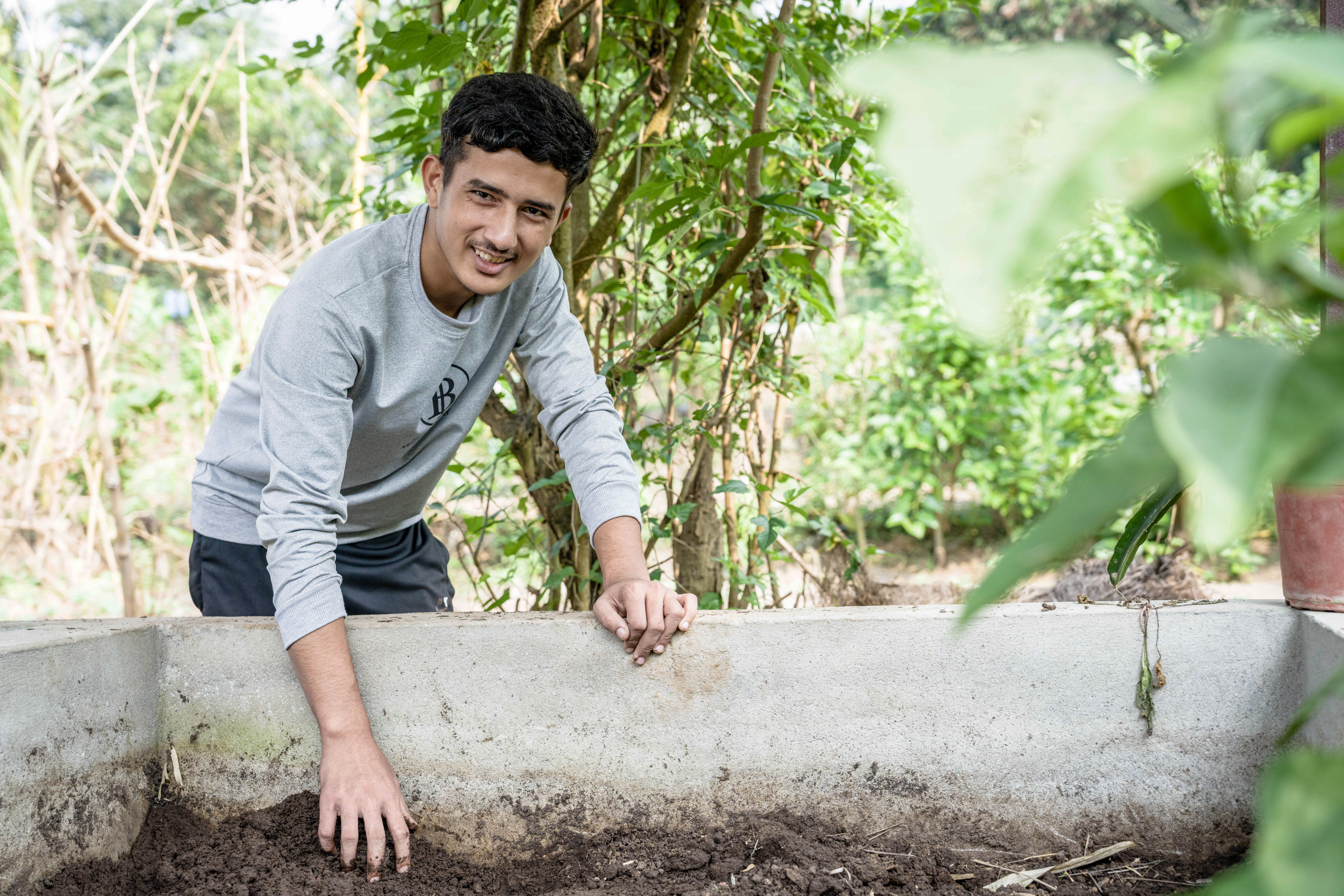 A young man in a gray long-sleeve shirt smiles while tending to soil in a raised garden bed surrounded by green plants and trees outdoors.