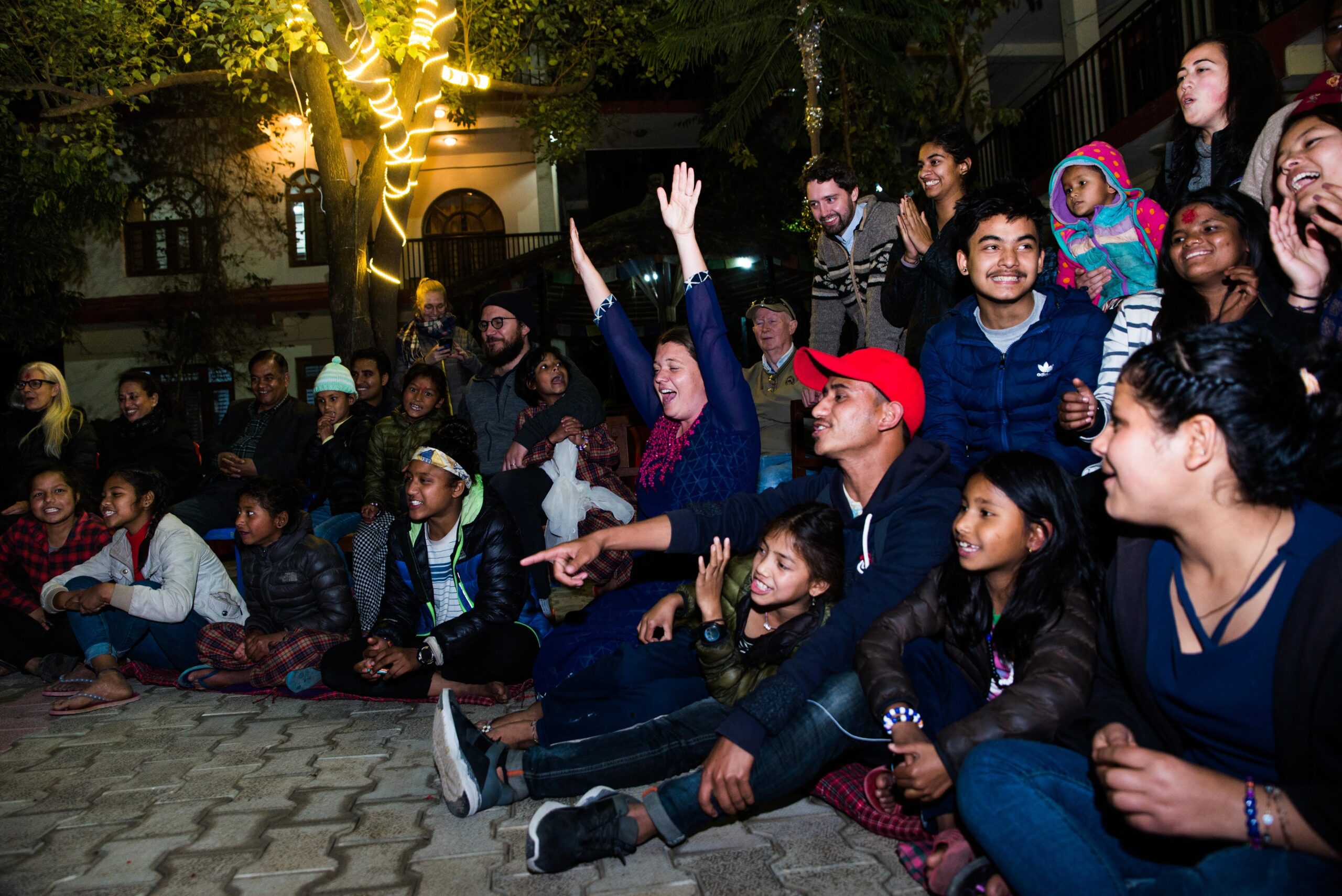 A diverse group of people, including children and adults, sit outdoors at night on cobblestone, smiling, laughing, raising hands, and watching something off-camera under trees decorated with string lights.