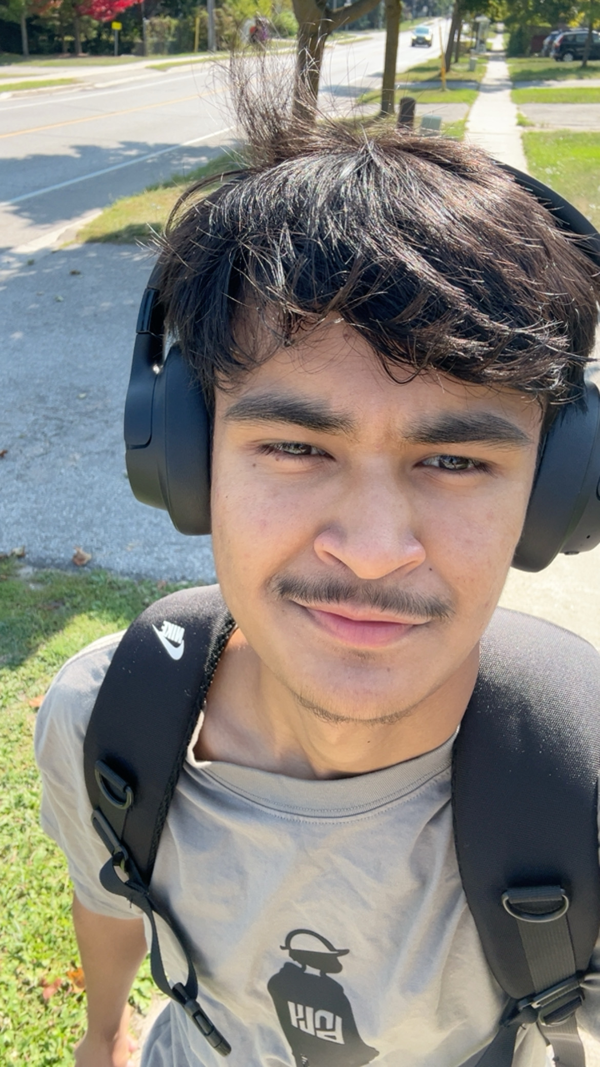 A young person with dark hair and headphones is wearing a beige shirt and a backpack, standing outside on a sunny day with grass and a sidewalk in the background.