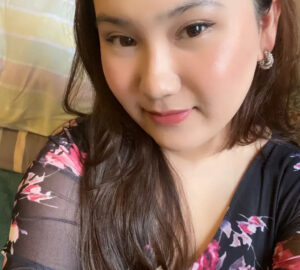 A woman with long brown hair is wearing a black top with pink floral patterns. She has light makeup, stud earrings, and is smiling softly while looking at the camera. The background shows a striped pillow or blanket.