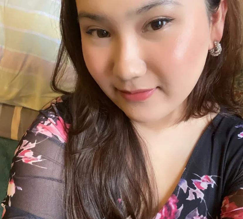 A woman with long brown hair is wearing a black top with pink floral patterns. She has light makeup, stud earrings, and is smiling softly while looking at the camera. The background shows a striped pillow or blanket.