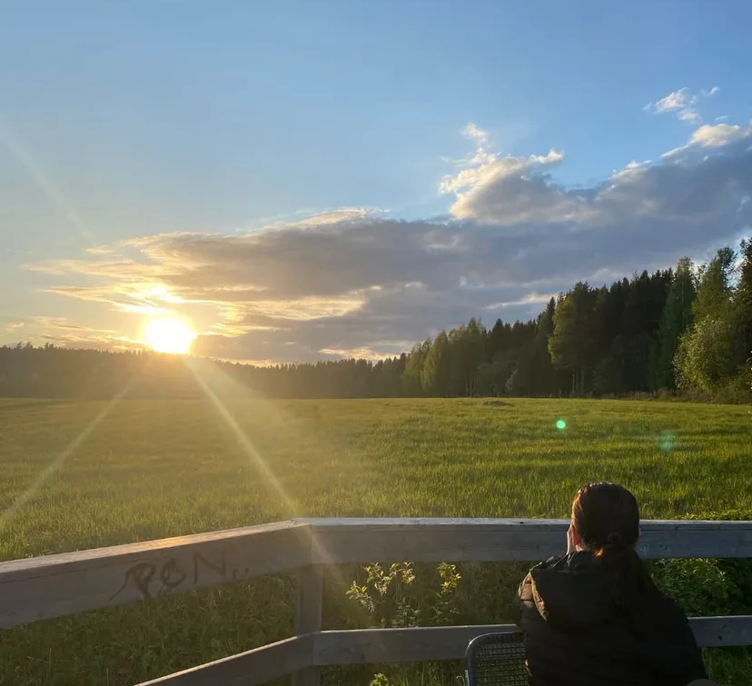 A person sits on a wooden bench facing a grassy field with trees, watching the sun set behind clouds in a clear blue sky. Sun rays stretch across the scene, creating a peaceful, serene atmosphere.