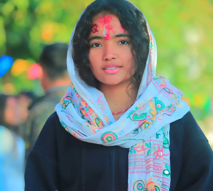 A young woman with curly hair wears a colorful, embroidered scarf over her head and shoulders and has red powder markings on her forehead. She stands outdoors with greenery and blurred people in the background.