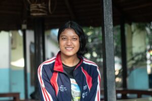 A young woman with dark hair smiles at the camera, wearing a navy blue and red jacket, standing in an outdoor shaded area with blurred background buildings and greenery.