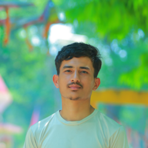 A young man with short dark hair and a mustache stands outdoors, wearing a light-colored t-shirt. The background is blurred with green trees and hints of colorful structures, suggesting a park or garden setting.