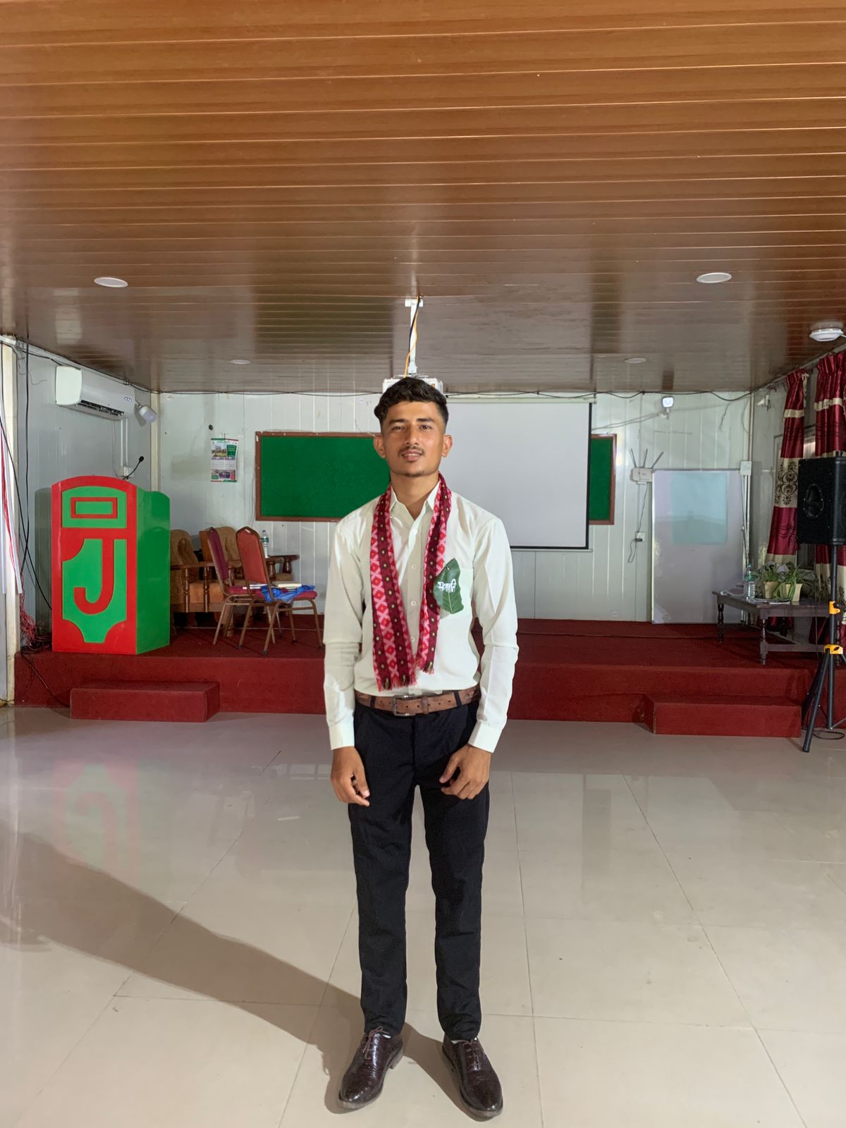 A young man in formal attire and a patterned scarf stands in a spacious room with a red stage, chairs, a green and red podium, and a projector screen in the background.
