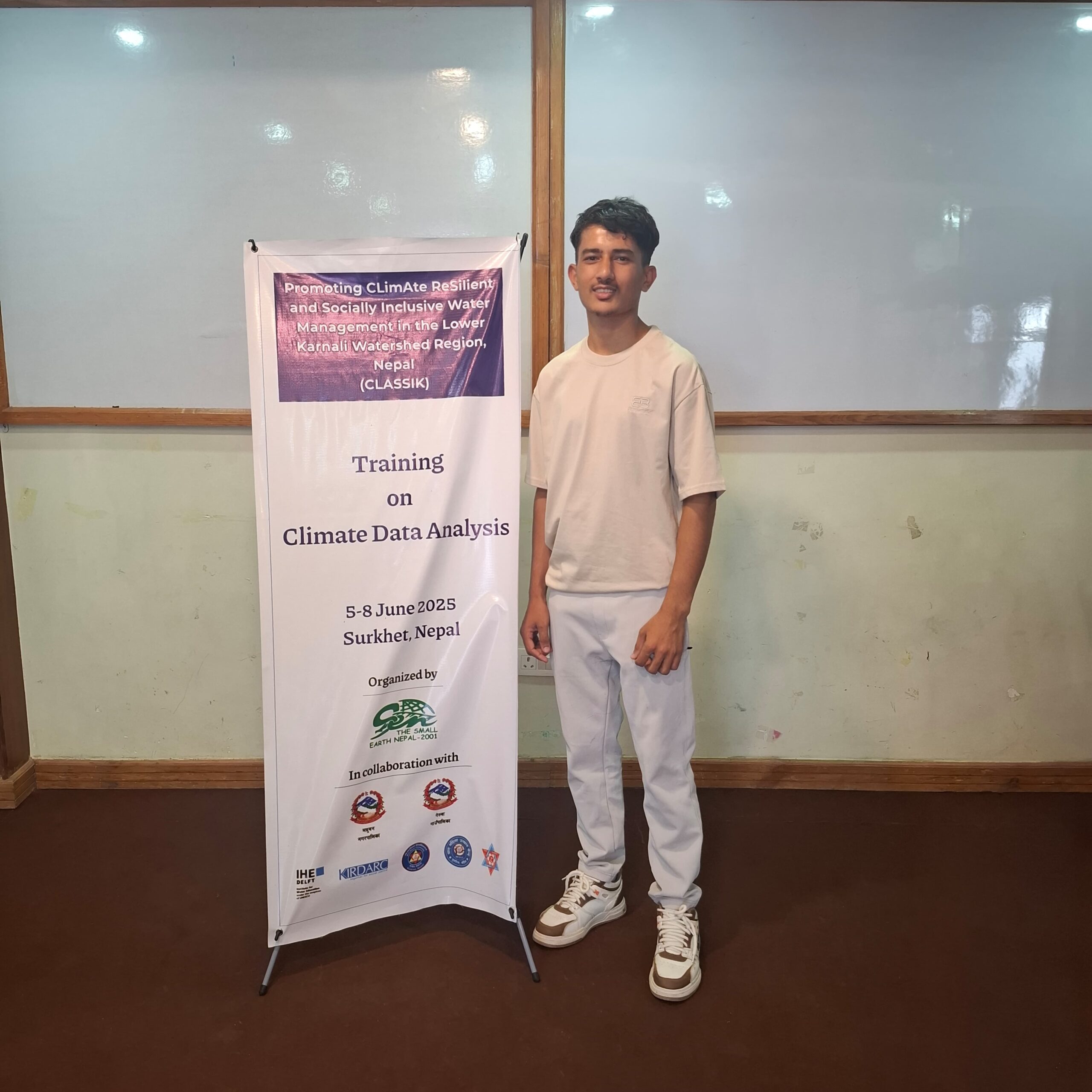 A young man stands indoors beside a roll-up banner for a "Training on Climate Data Analysis," held June 5-8, 2025, in Surkhet, Nepal. The banner displays event details and partner logos.