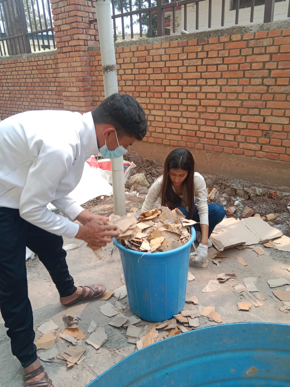 A man and a woman are sorting and placing pieces of cardboard into a large blue bin outdoors near a brick wall. The man is wearing a face mask and bending over, while the woman is crouching and wearing gloves.