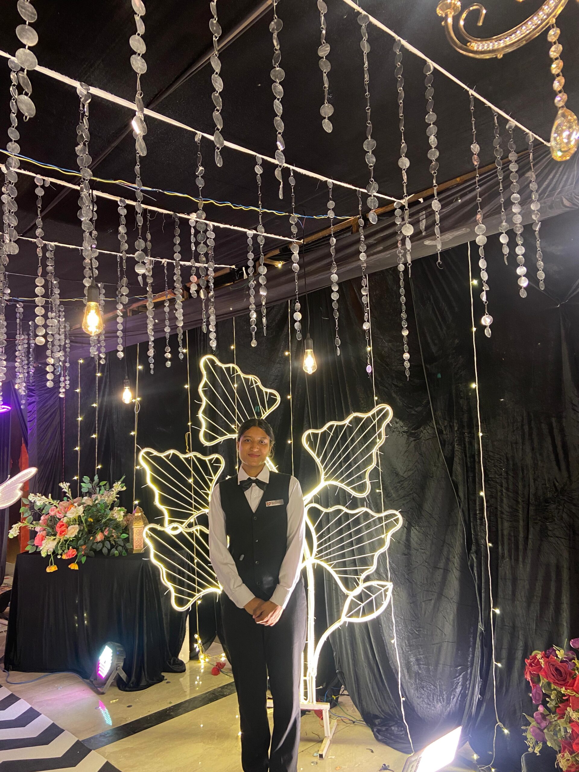 A smiling person in a black vest and white shirt stands indoors under hanging crystal decorations, with illuminated leaf-shaped lights and flower arrangements in the background.