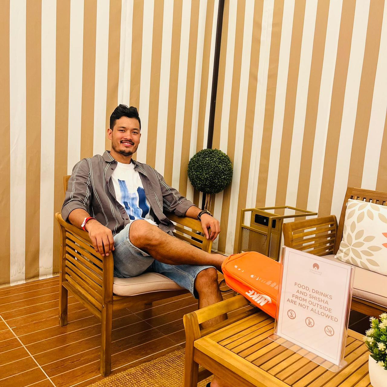 A man sits smiling on a wooden chair in a room with beige and white striped walls. He wears casual clothes and has one leg crossed. A sign on the table says food, drinks, and snacks from outside are not allowed.