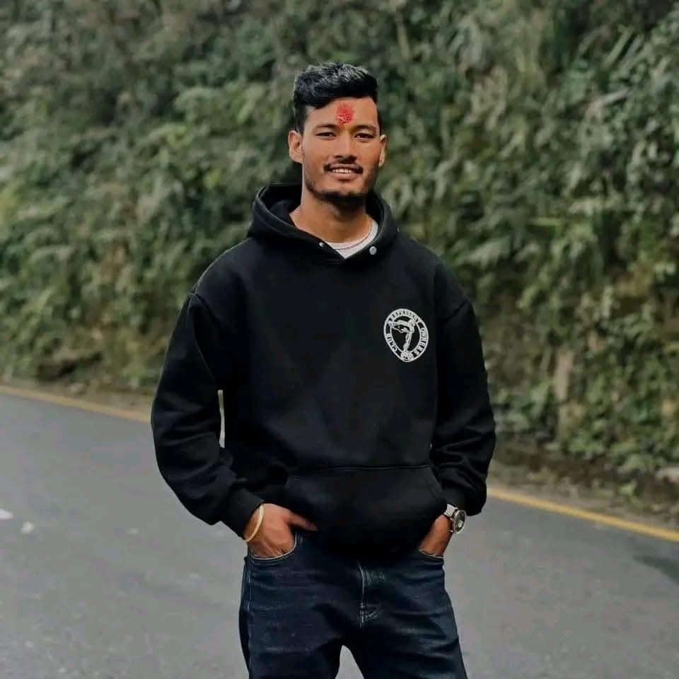 A man with a red mark on his forehead stands on a roadside with greenery in the background, wearing a black hoodie and jeans, with his hands in his pockets and smiling at the camera.