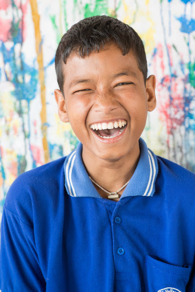 A boy wearing a bright blue collared shirt laughs joyfully, showing his teeth, with a colorful, paint-splattered wall in the background.