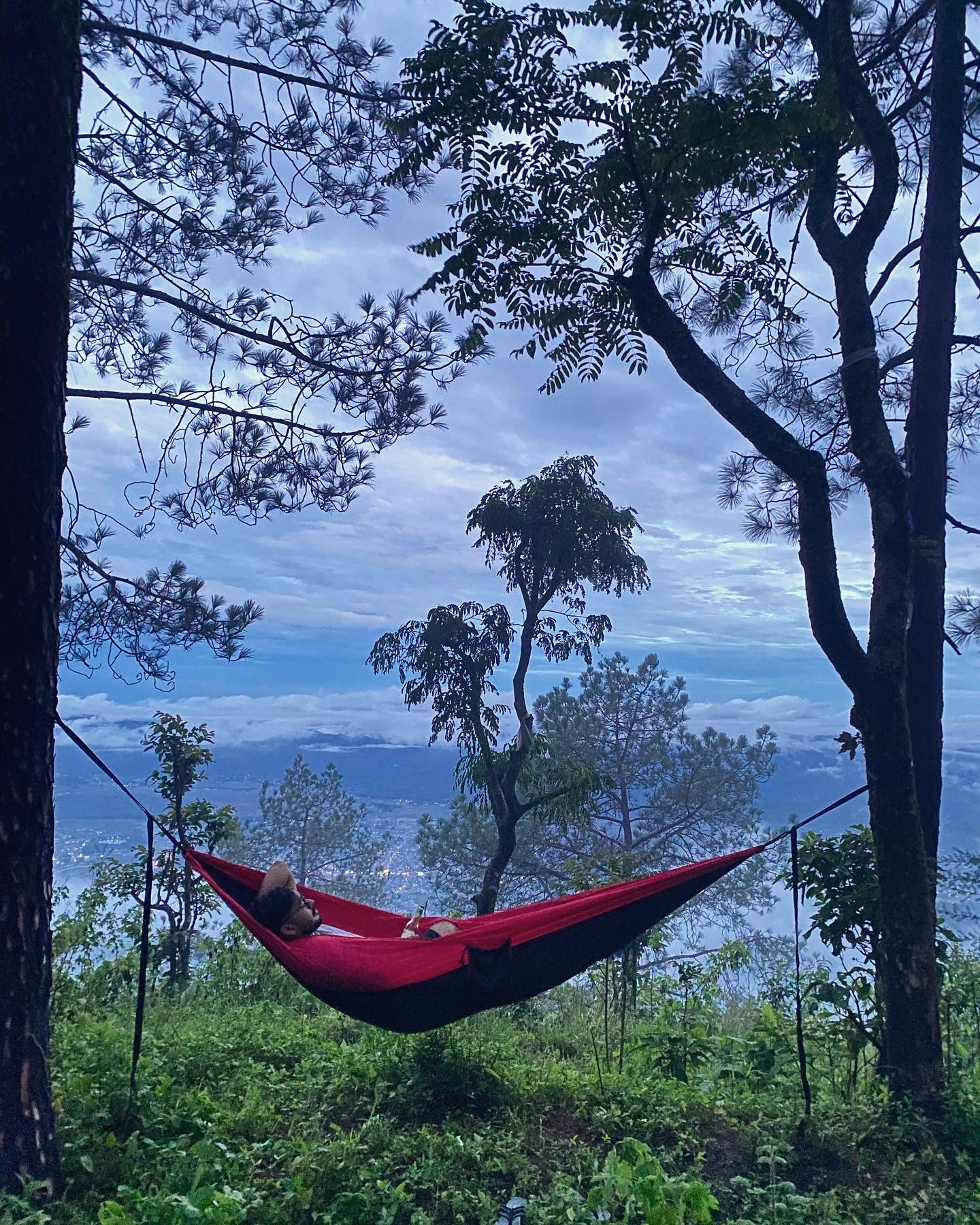 A person relaxes in a red hammock tied between two trees, surrounded by greenery, with a scenic mountain and cloudy sky in the background.