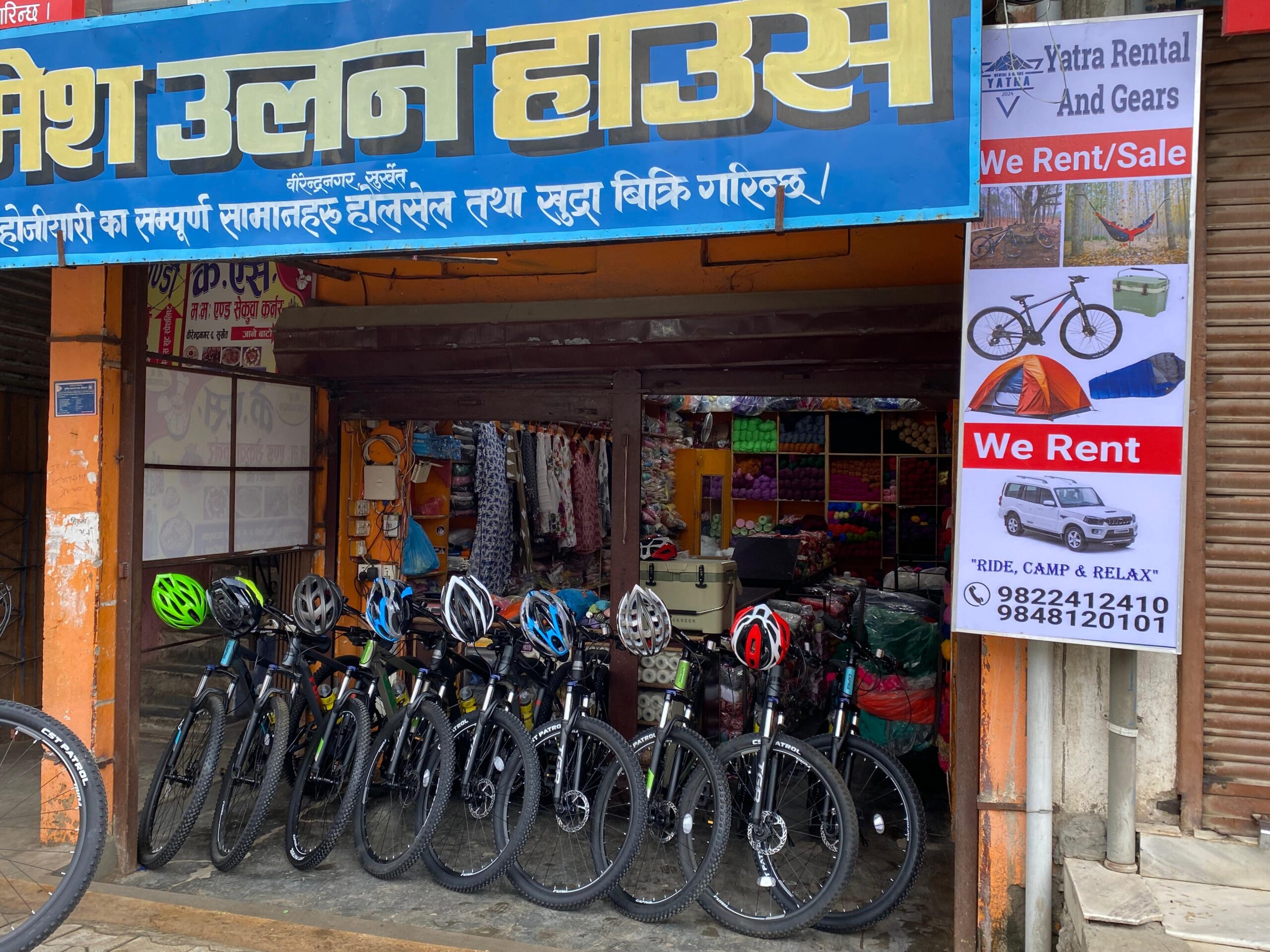 Row of bicycles with helmets parked outside a rental shop in India. Signboards above and beside the shop advertise camping gear, bicycles, car rentals, and contact numbers. Shop entrance is open with items visible inside.