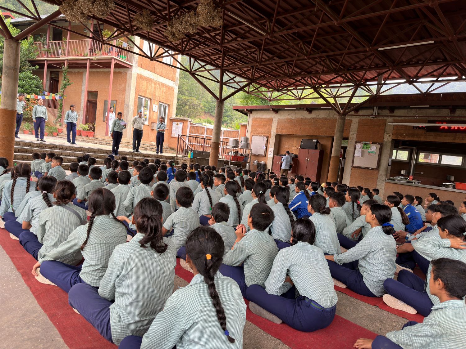 A large group of students in uniform sit cross-legged on the floor inside an open-air, covered area, attentively facing a group of teachers or speakers standing on a stage at the front.