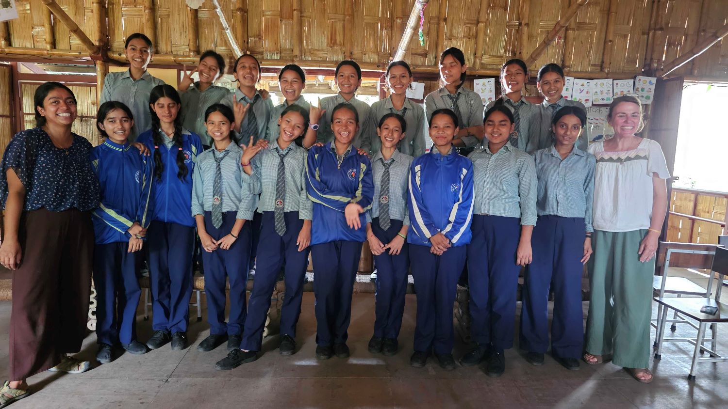 A group of smiling students in school uniforms and two adults stand together inside a classroom with bamboo walls, posing for a group photo. Some students wear blue tracksuits, while others wear green shirts and ties.