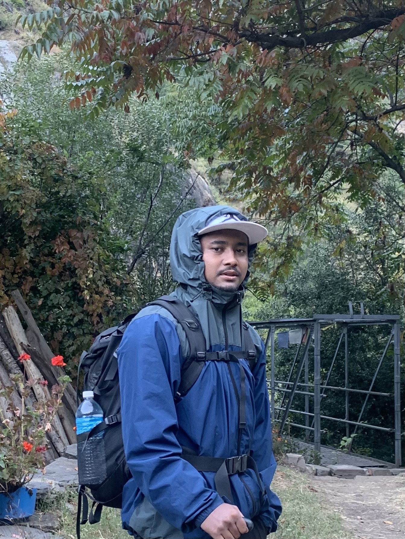 A person wearing a blue jacket, backpack, and cap stands outdoors near greenery, flowers, and a metal bridge, with trees and a hillside in the background.