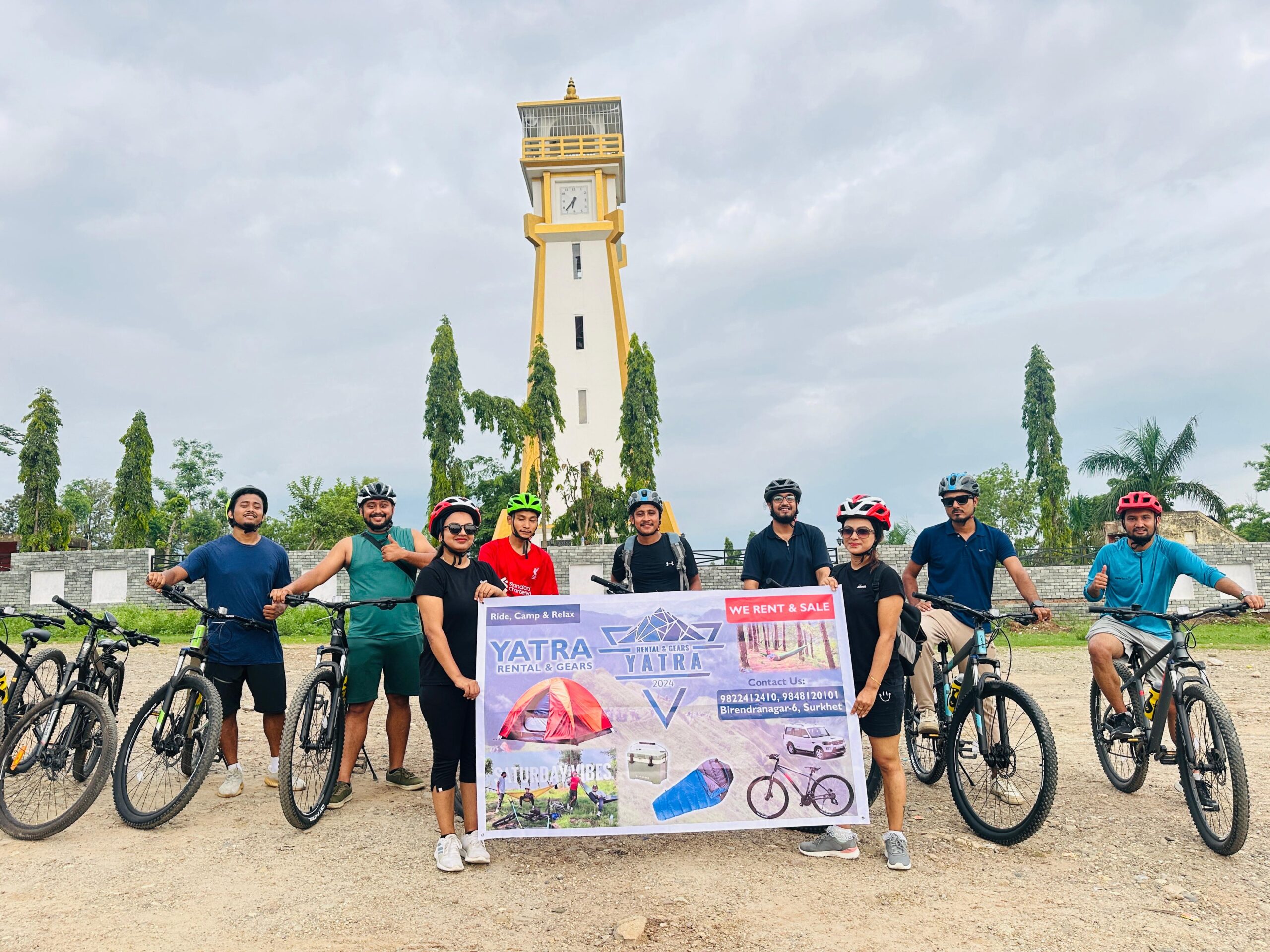 A group of cyclists with helmets poses with their bikes and a promotional banner in front of a tall clock tower, surrounded by greenery and trees on a cloudy day.