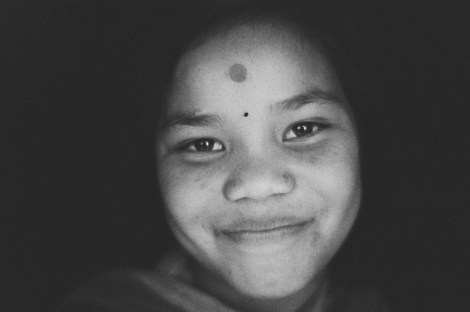 Black and white close-up of a young person smiling softly, with a bindi and a dot on their forehead, set against a dark background.