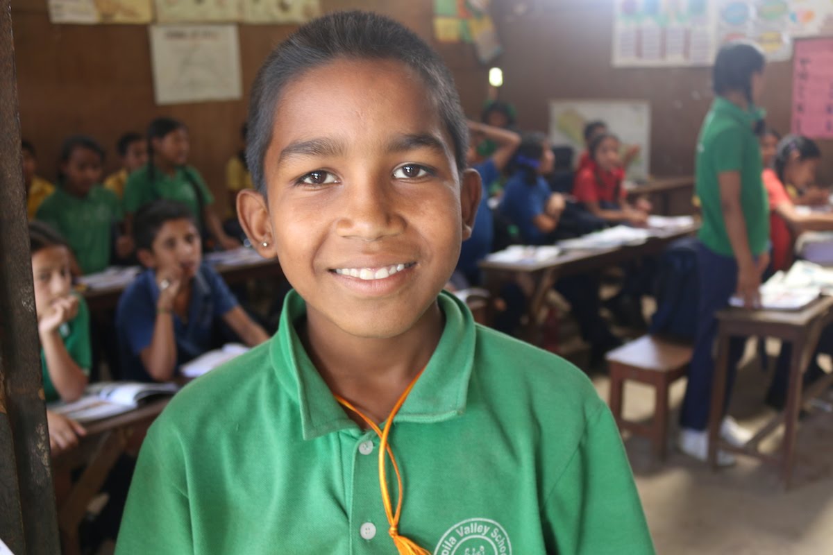A young student wearing a green shirt smiles at the camera inside a classroom, while other students sit at desks and study in the background. The classroom looks bright and active.
