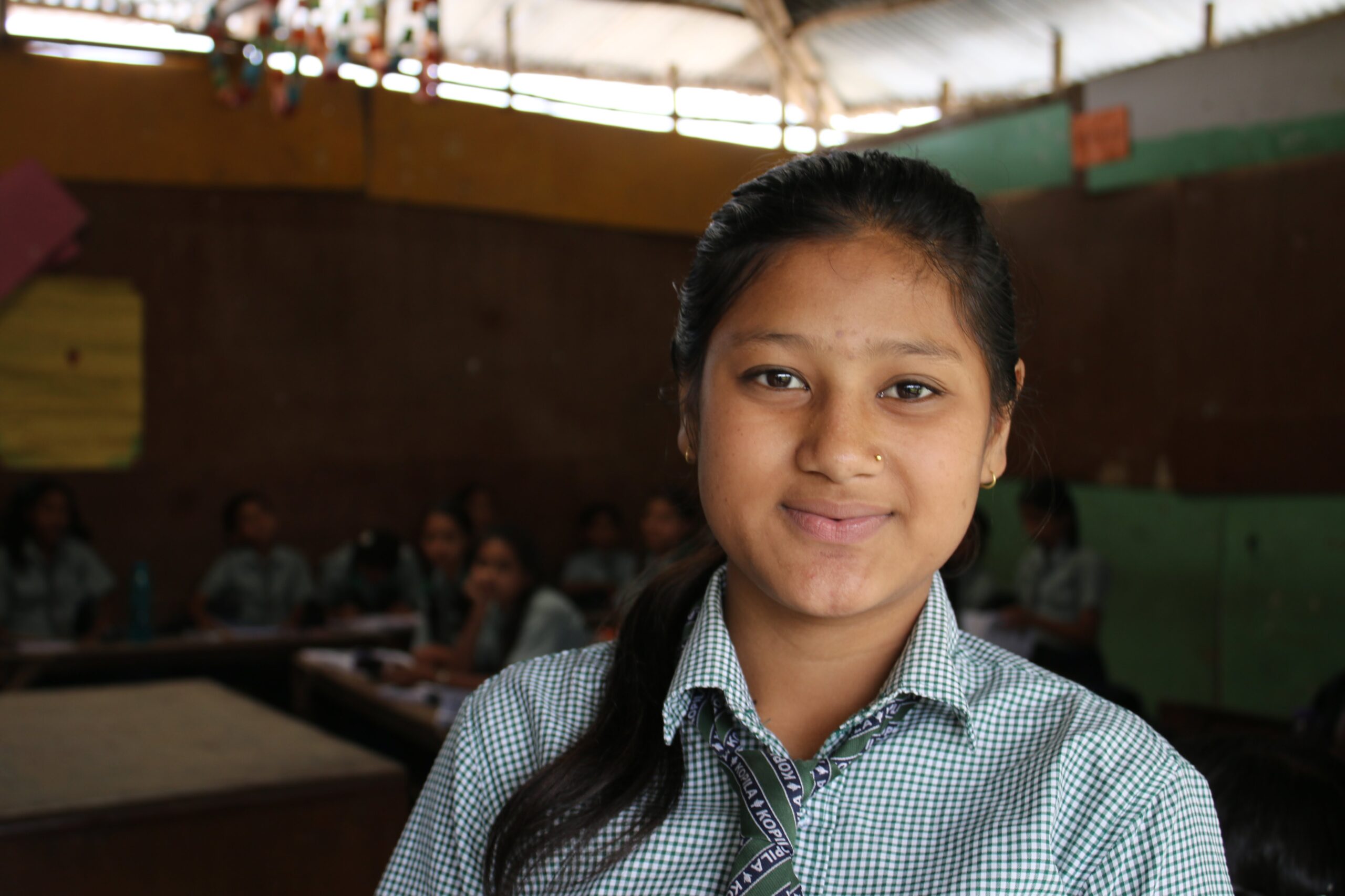A girl in a school uniform smiles at the camera in a classroom, with other students seated in the background. The classroom has brown walls and a corrugated metal roof.