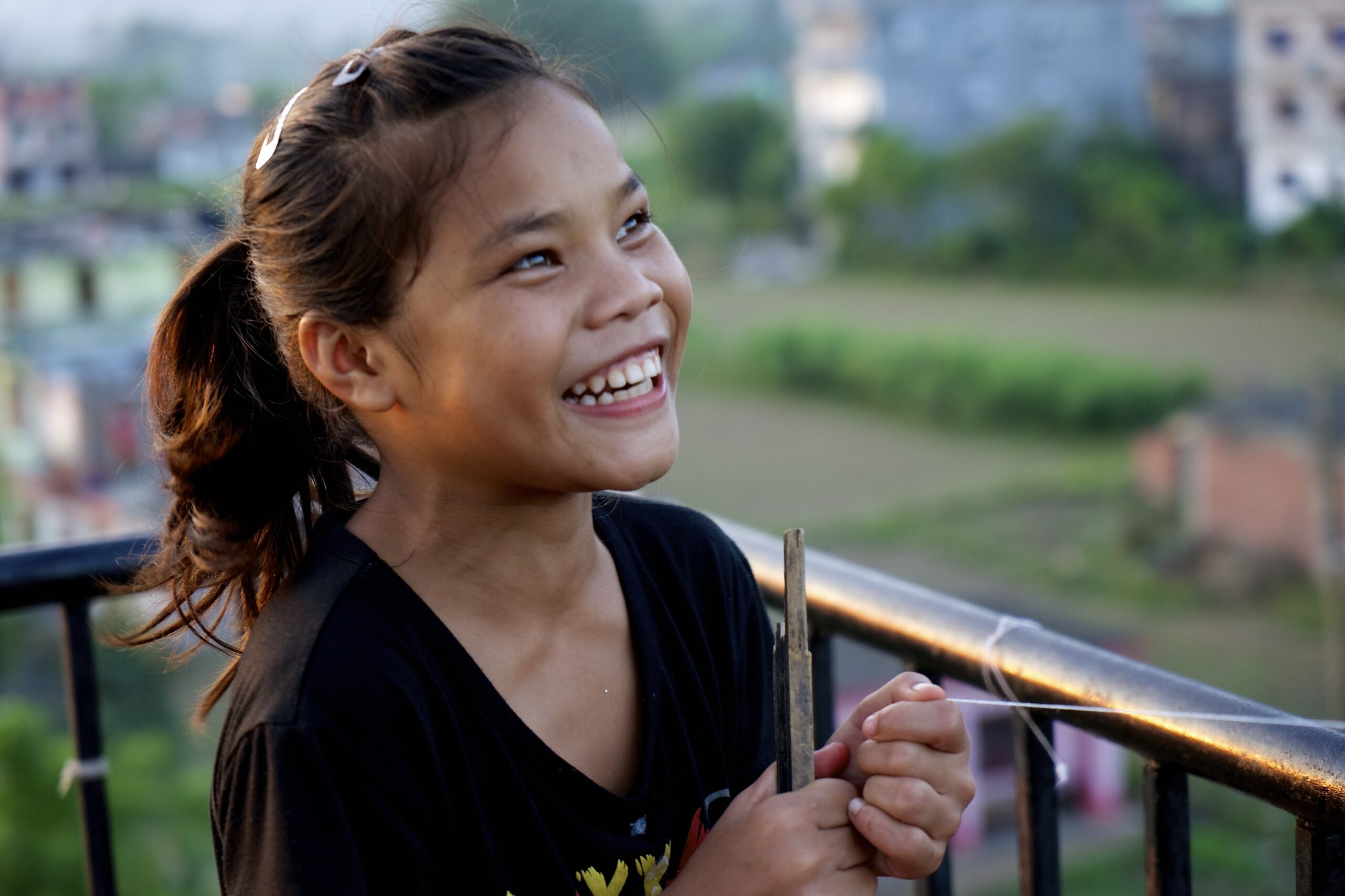 A young girl with brown hair in a ponytail smiles joyfully while standing outdoors by a metal railing, holding a stick, with a blurred background of green fields and buildings.