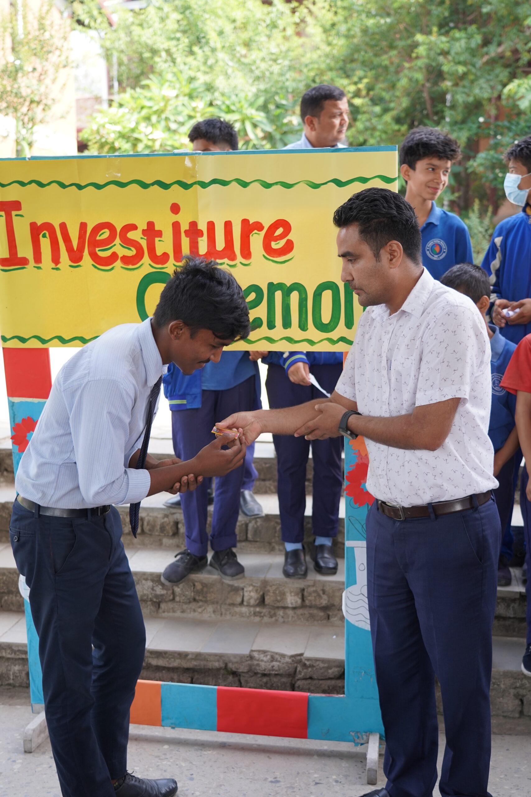 A student in uniform bows while receiving a badge from a man during an investiture ceremony. Several students in blue uniforms stand in the background near a colorful sign.