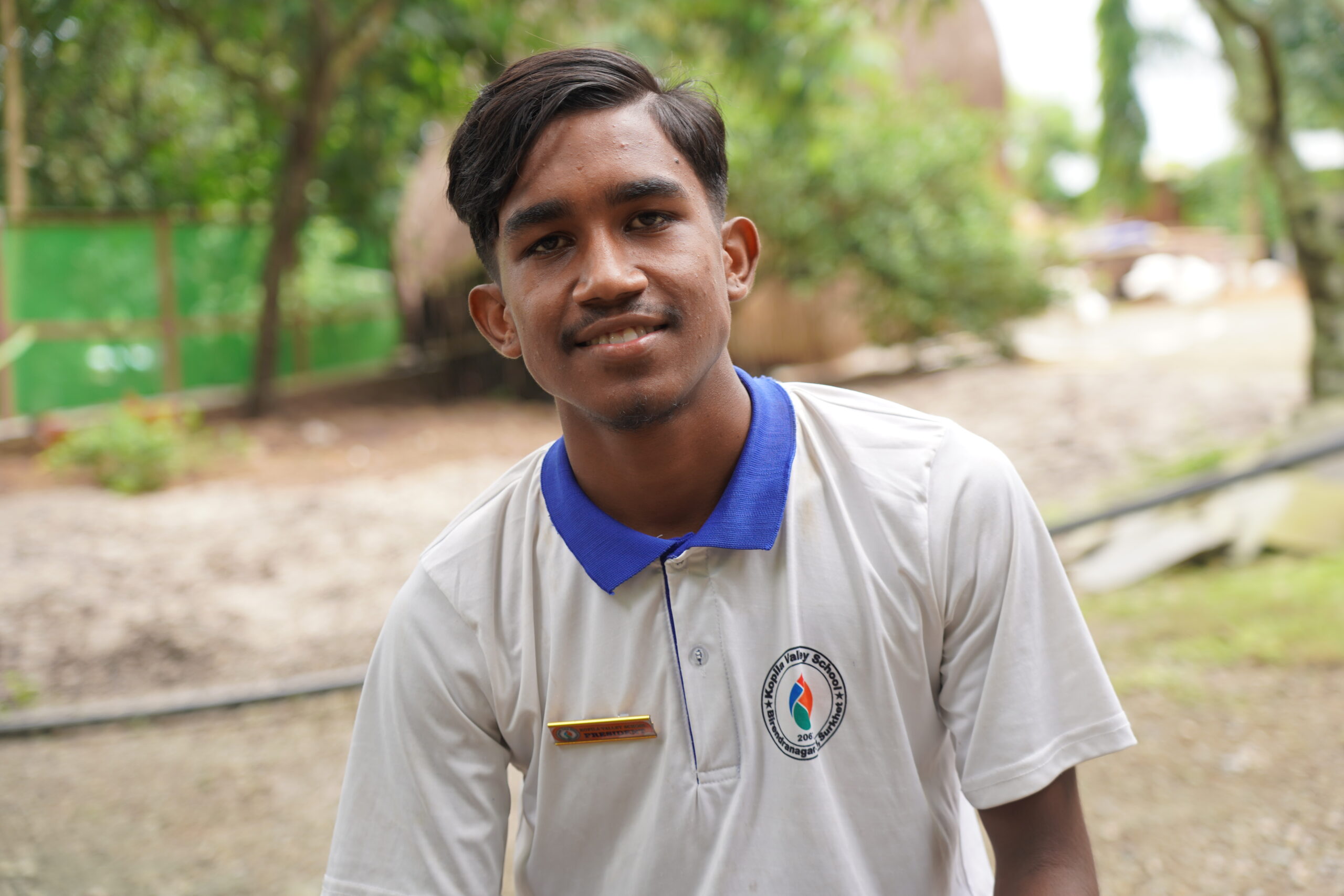A young man with short dark hair, wearing a white collared shirt with a blue trim and an emblem, smiles at the camera outdoors with greenery in the background.