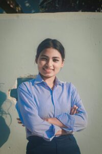 A young woman with dark hair, wearing a light blue button-up shirt, stands in front of a pale wall with her arms crossed, smiling confidently at the camera. Sunlight casts soft shadows on the wall behind her.
