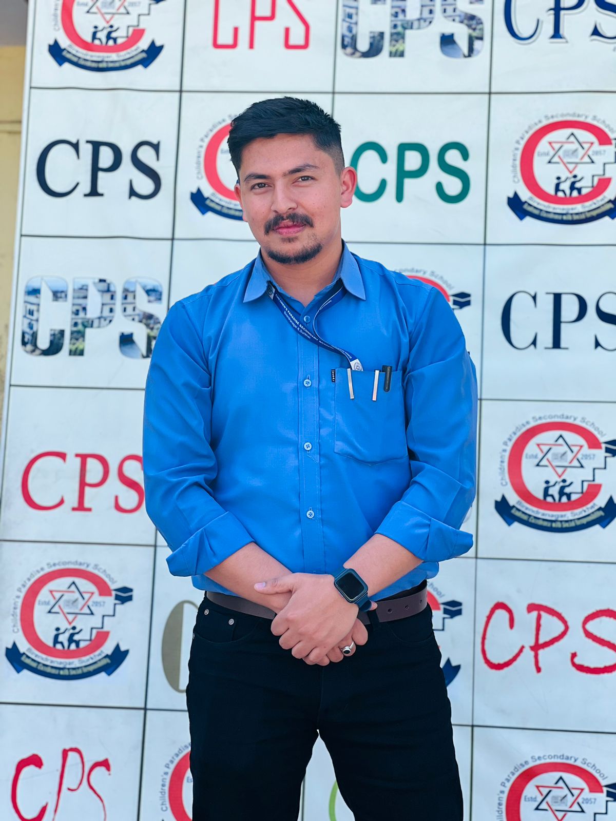 A man with short dark hair and a trimmed beard stands in front of a backdrop with various CPS and KK logos. He is wearing a blue button-up shirt, black pants, and a smart watch, with pens in his shirt pocket.