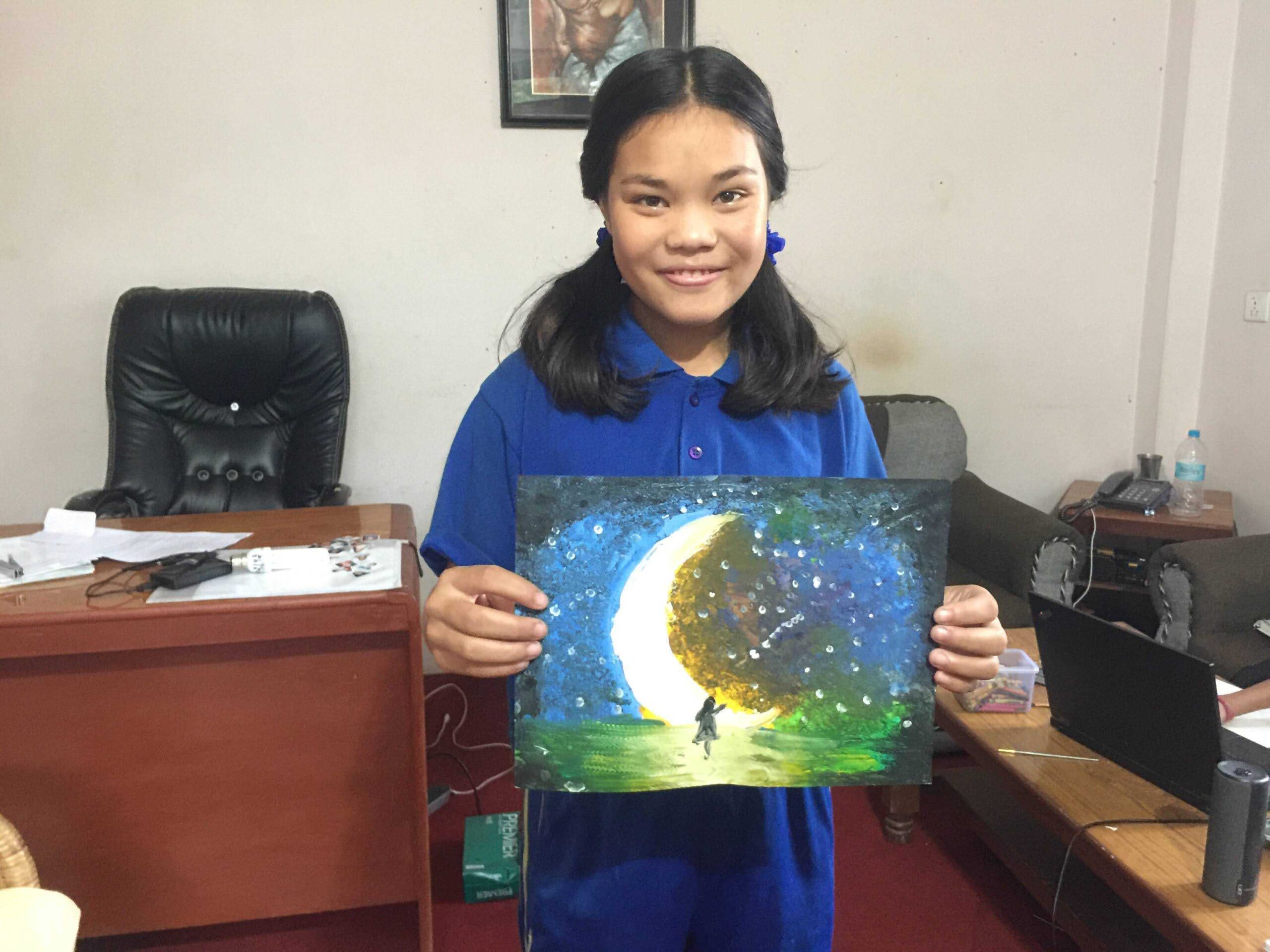 A girl in a blue shirt smiles and holds a painting of a crescent moon with a starry sky and a figure standing beneath it. She stands in an office with a desk, chair, and scattered papers in the background.