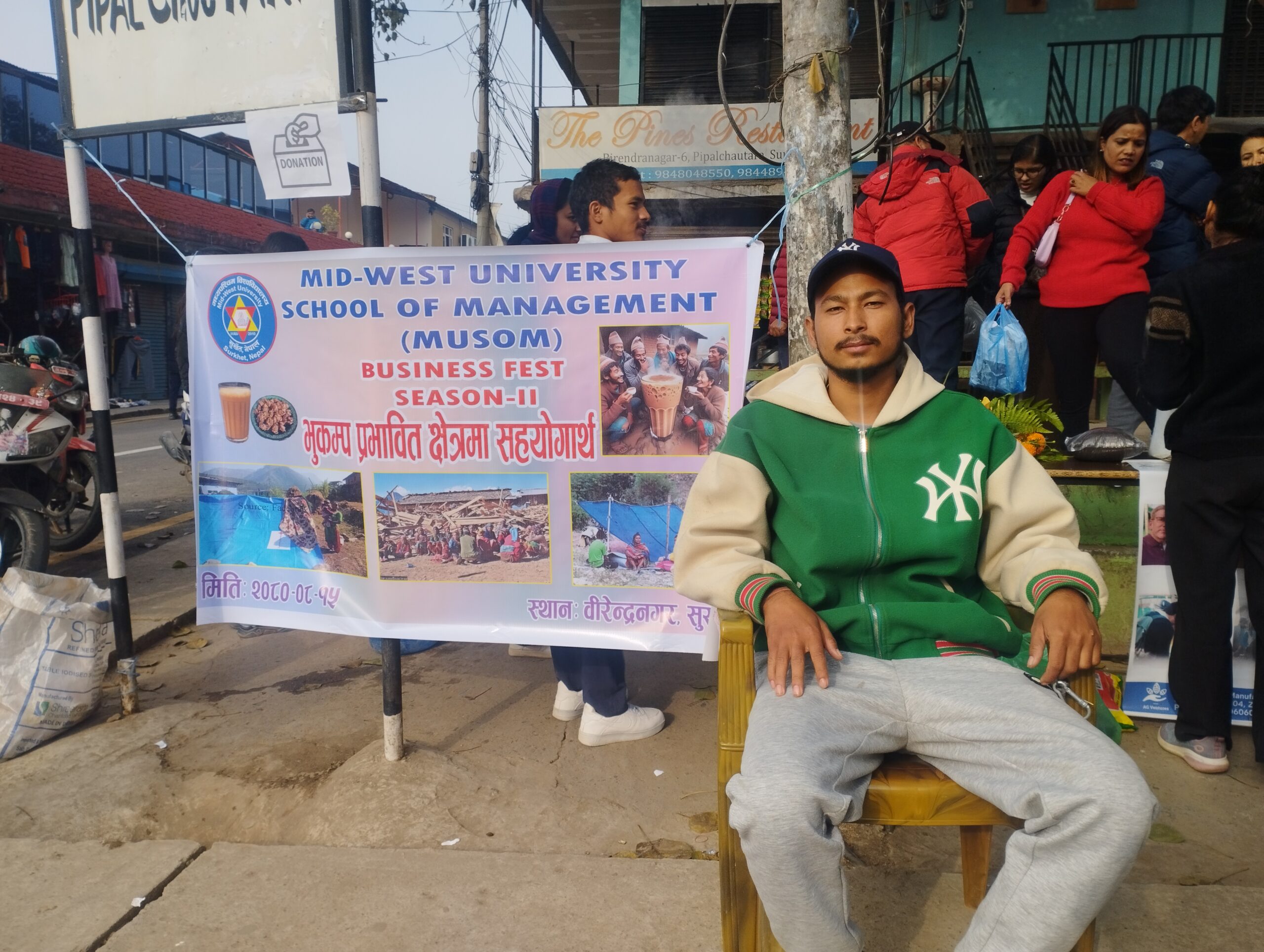 A man in a green jacket and cap sits on a chair in front of a "Mid-West University School of Management Business Fest Season-II" banner, with people gathered and shops visible in the background.