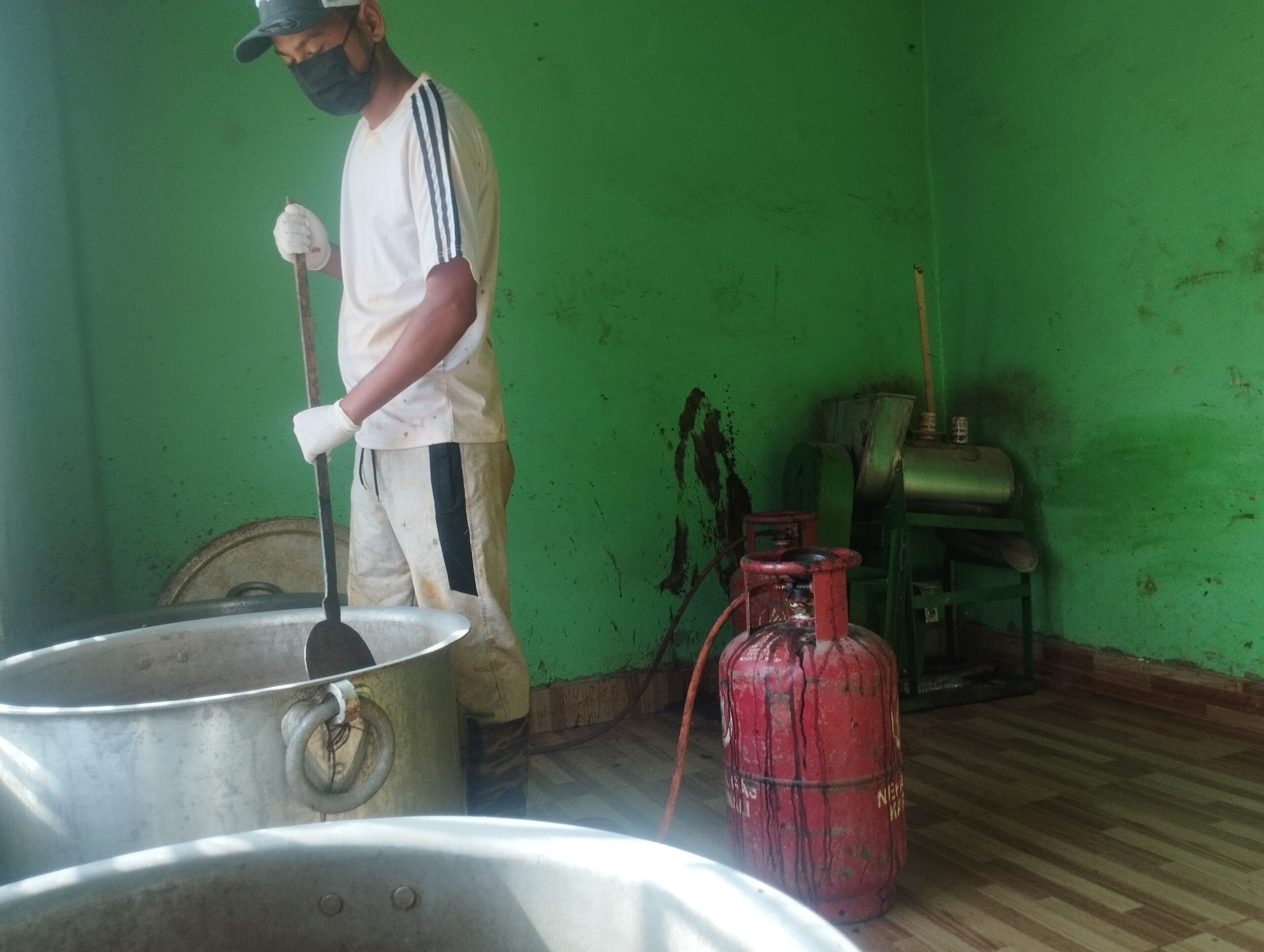 A person wearing a mask, cap, gloves, and casual clothes stirs a large metal pot in a green-walled room. A red gas cylinder and a metal machine are nearby on the wooden floor.