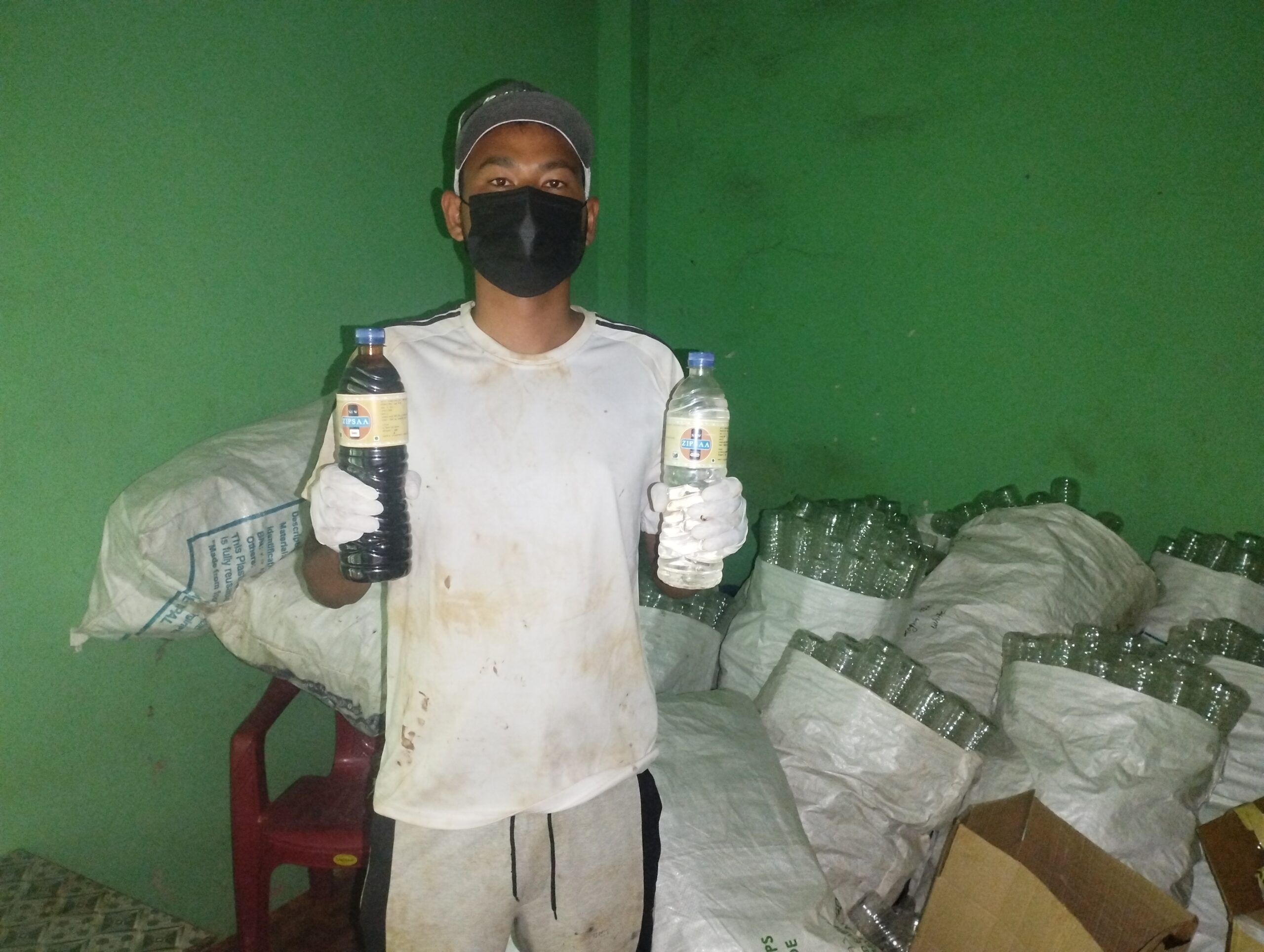 A person wearing a cap, face mask, white gloves, and a stained white shirt holds a dark bottle in one hand and a clear bottle in the other. They stand in front of bags and stacks of empty plastic bottles.