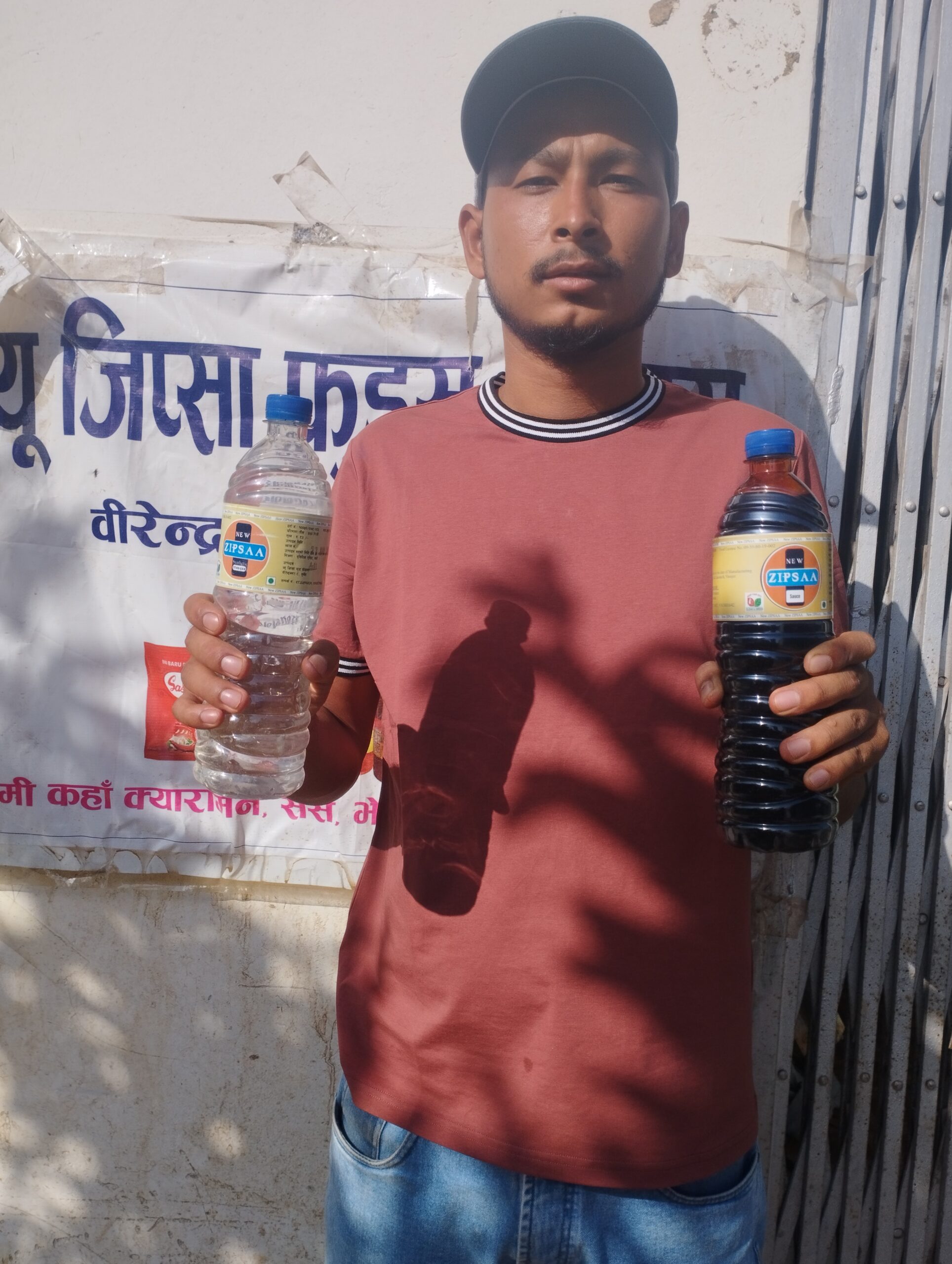 A man in a red shirt and cap stands outdoors, holding a clear bottle with light liquid in his left hand and a similar bottle with dark liquid in his right hand, against a wall with a partially visible sign.