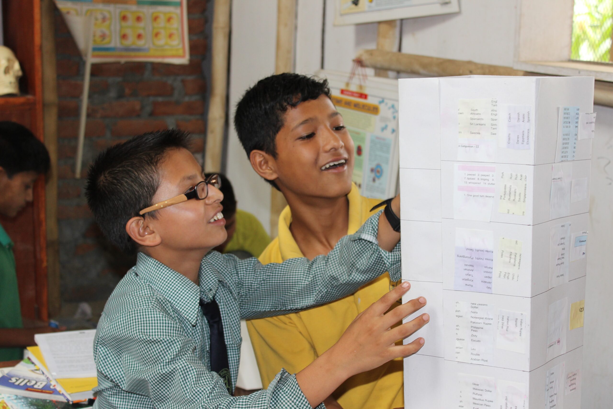 Two boys, one wearing glasses and a tie, and the other in a yellow shirt, smile and stack paper blocks with printed lists in a classroom setting. Books, posters, and another student are visible in the background.