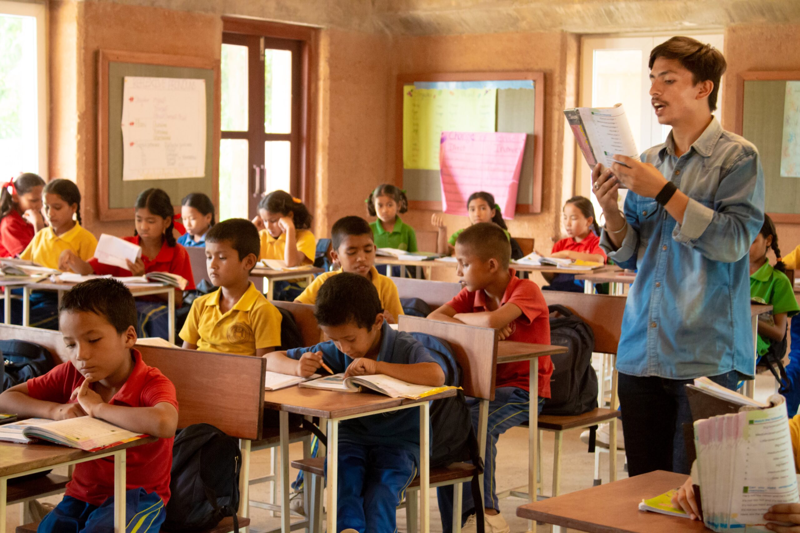 A teacher stands at the front of a classroom reading from a book, while students in colorful uniforms sit at desks, writing and paying attention to the lesson. Posters and windows are visible in the background.