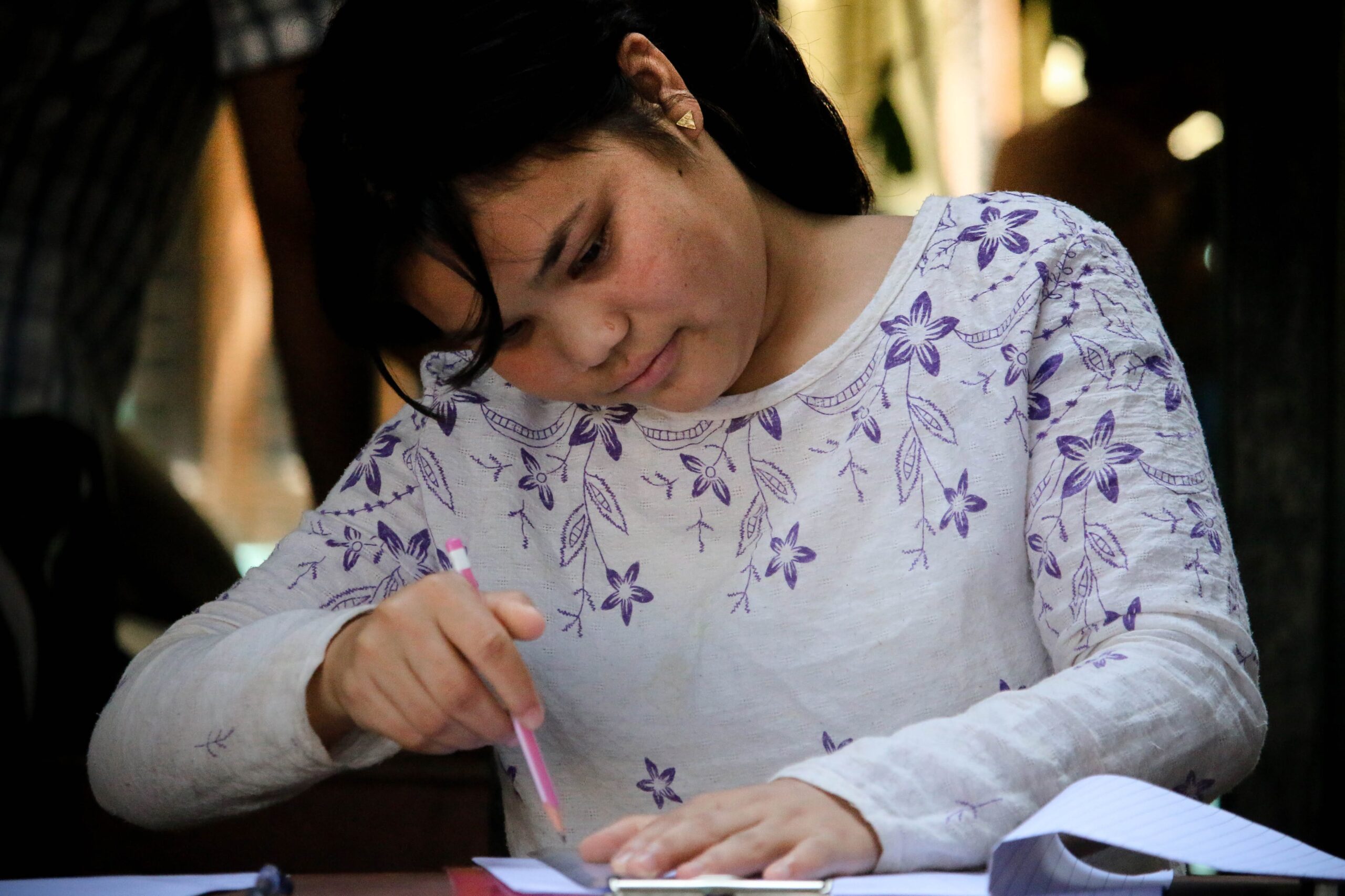 A young woman with dark hair, wearing a white shirt with purple floral patterns, is focused on writing in a notebook with a pink pencil at a table.