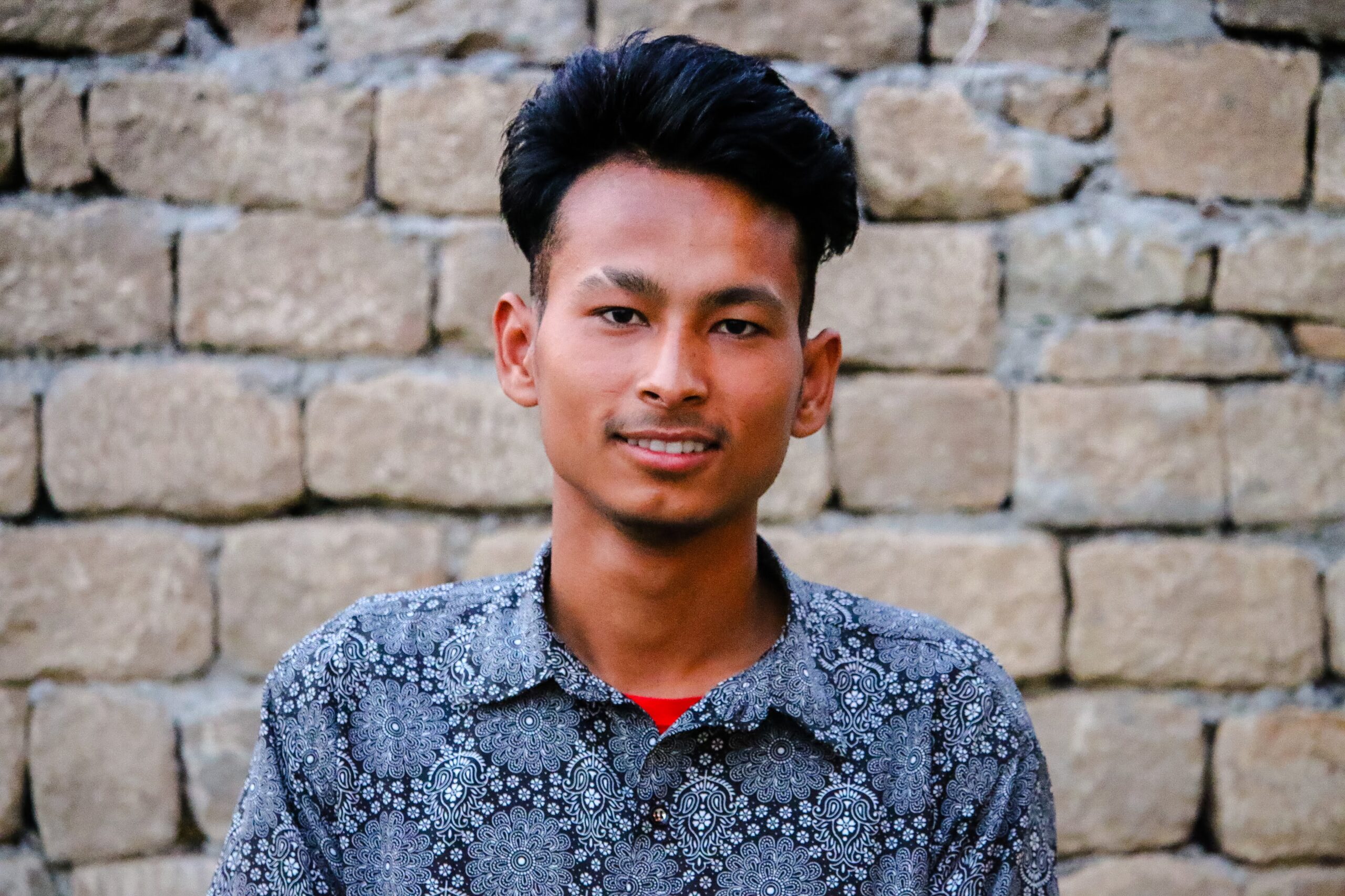 A young man with dark hair wearing a patterned shirt stands in front of a stone brick wall, smiling slightly at the camera.