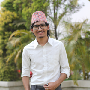 A man wearing glasses, a white shirt, and a colorful patterned topi stands outdoors, smiling, with blurred green trees and plants in the background.