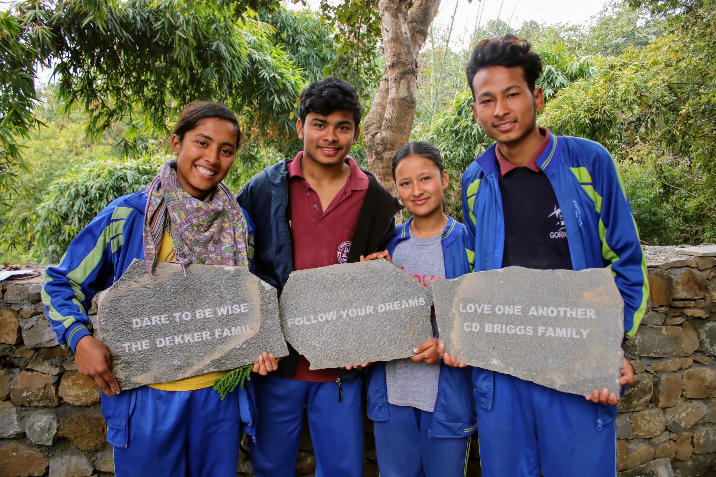 Four young people stand in front of a stone wall outdoors, each holding engraved stones with inspirational messages about wisdom, dreams, and love from different families. They are smiling and dressed in casual clothing.