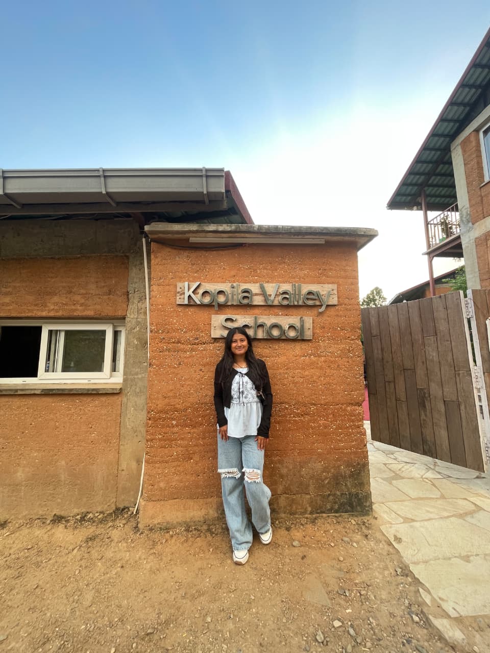 A young woman stands smiling in front of a brick wall with a sign that reads "Kopila Valley School." She is wearing a black jacket, light blue shirt, and ripped jeans. The sky is clear and buildings surround the area.