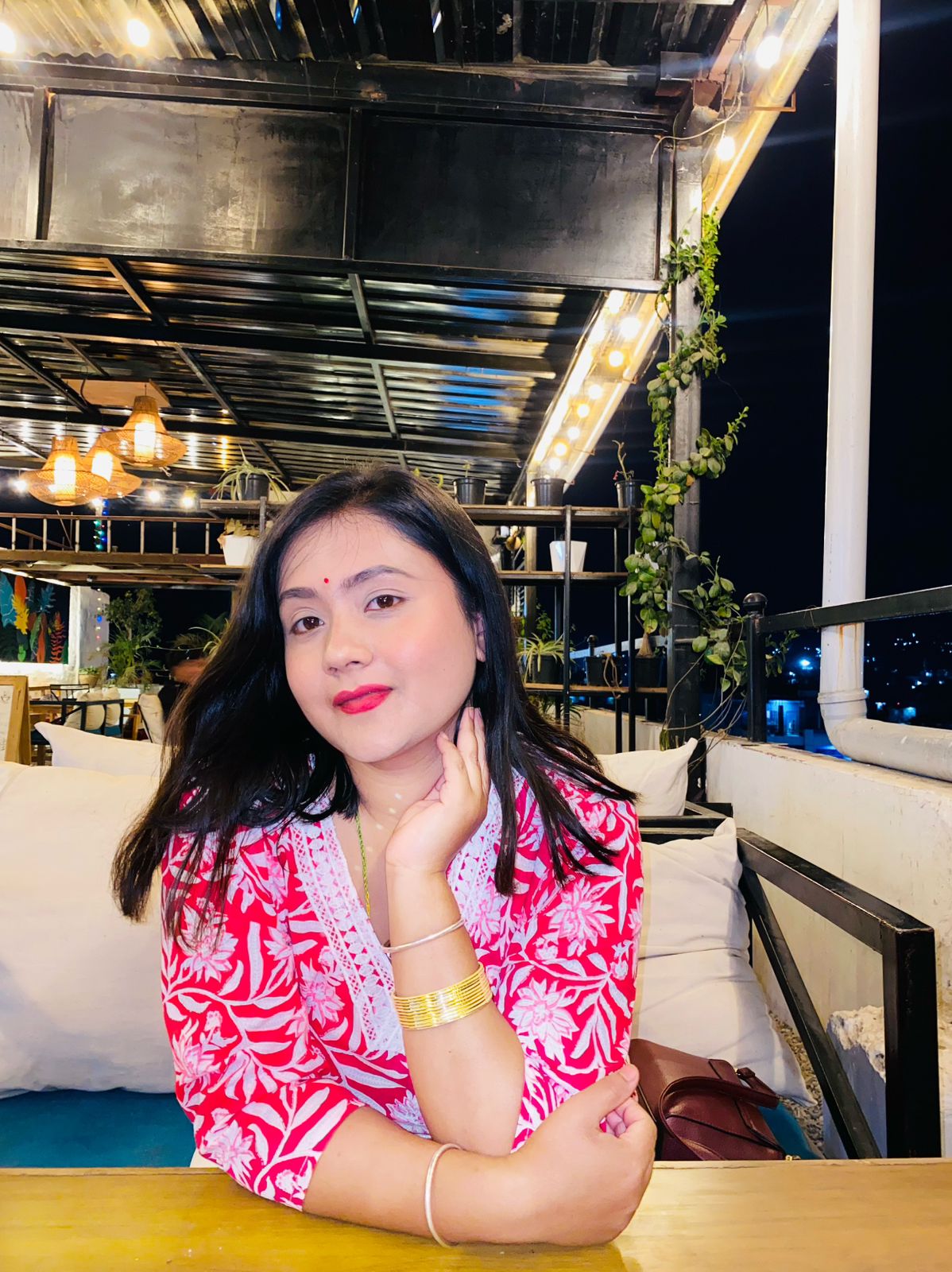 A woman with long dark hair, wearing a red and white traditional outfit, gold bangles, and red lipstick, sits at a wooden table in a well-lit outdoor restaurant at night. She is smiling softly at the camera.