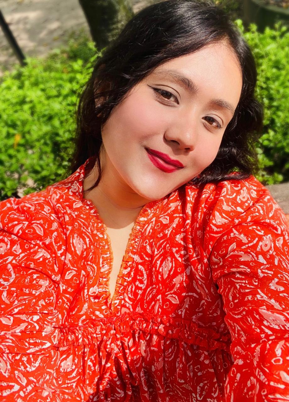 A person with long dark hair wearing a bright red patterned top smiles while taking a selfie outdoors. Green bushes and sunlight are visible in the background.