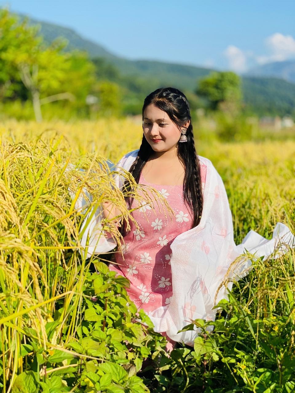 A woman in a pink floral dress and white shawl stands in a lush green field with tall grass and crops, with mountains and trees in the background under a bright blue sky.