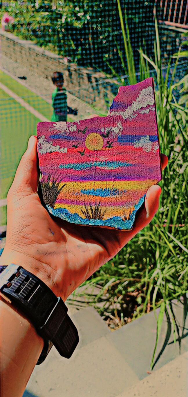 A hand holds a painted rock featuring a colorful sunset, grass, and clouds. In the blurred background, a child stands on green grass near a stone wall and tall plants.