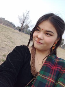 A woman wearing traditional jewelry, a red bindi, and a colorful plaid saree takes a selfie outdoors on a grassy field with bare trees in the background.