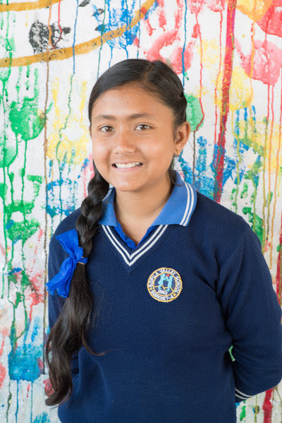 A girl with long, braided hair wearing a dark blue school sweater and collared shirt stands smiling in front of a colorful, paint-splattered wall.