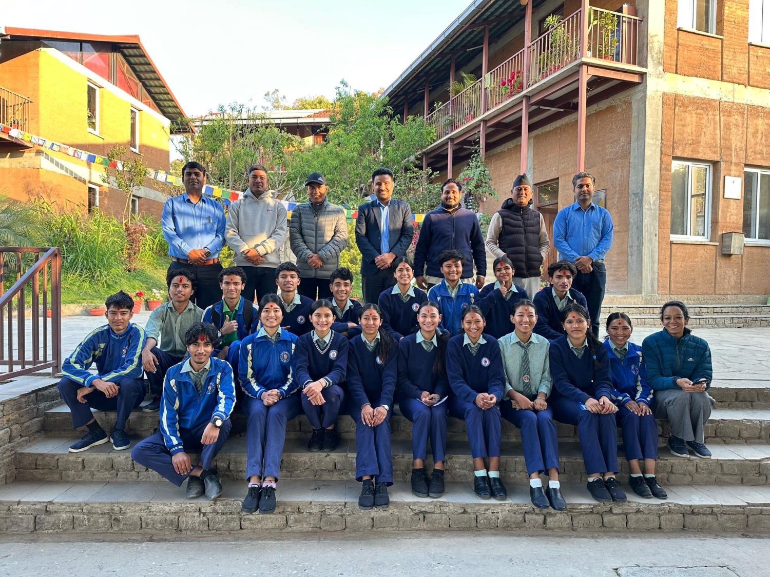A group of students in school uniforms and teachers in casual attire pose for a photo on steps outside a school building, with greenery and buildings in the background.