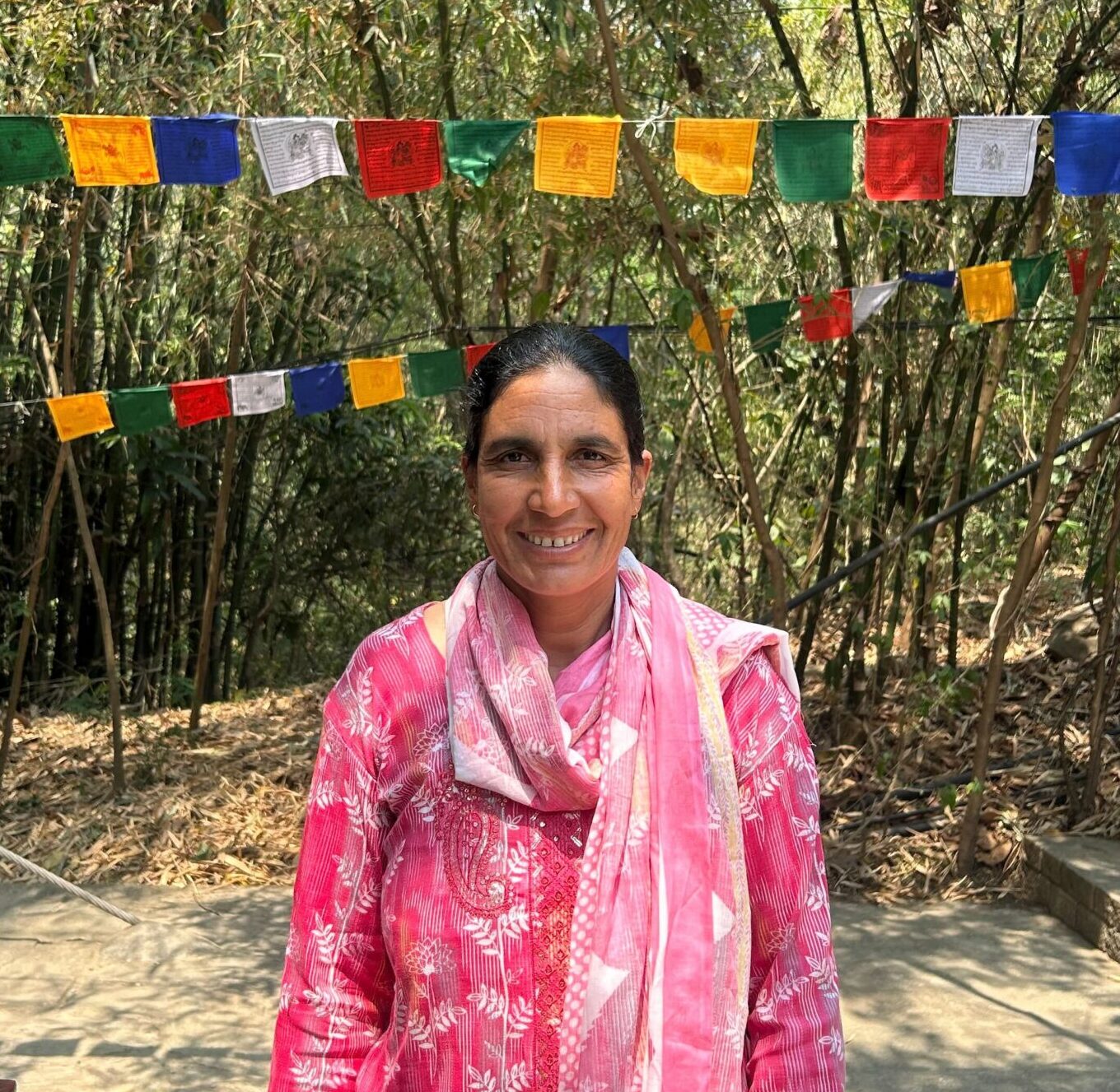 A woman in a pink patterned dress and scarf stands smiling outdoors in front of colorful prayer flags and bamboo trees on a sunny day.
